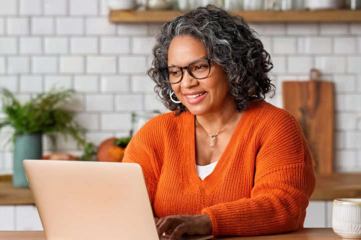 Middle Aged Woman Using Laptop In Kitchen On Table Wearing A Big Orange Jumper Aspect Ratio 720 480