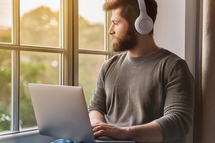 Young White Man With Beard Next To A Window With Headphones On Using A Laptop Aspect Ratio 720 480